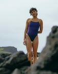 JAYD Swimwear model Sophie in a purple acai Lennox one piece swimsuit standing on rocky terrain with a natural landscape background