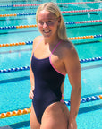 JAYD athlete Ella Ramsay wearing a dark acai Lennox one-piece swimsuit with bright fairy floss pink trim, stands in front of an outdoor swimming pool with lane dividers.