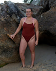 Athlete Kim Doyle standing on a beach in front of a collection of rocks, wearing the JAYD Swimwear cinnamon one piece with contrasting pink fairy floss straps.