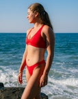 JAYD model wearing the bright ruby red Amalfi bikini bottoms with matching Vienna top, stands on a rocky shoreline with ocean waves and a clear blue sky behind her.