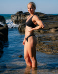 JAYD athlete Dom wearing the black Amalfi bikini bottoms and matching Vienna top, stands in shallow ocean water on a rocky beach, with waves and rocks visible in the background.