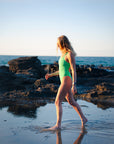 JAYD model woman wearing the green meadow one-piece Apsley swimsuit, walking through shallow water on a rocky beach with the ocean and clear sky in the background.