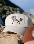 Cream JAYD Swimwear baseball hat with brown palm trees & swimsuit embroidery sitting on a rock.