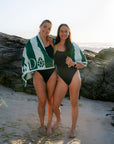 JAYD models Jenaya and Emily standing on a sandy beach with rocks behind them, wearing black one-piece swimsuits and wrapped in green and white towels with a visible 'JAYD' logo.