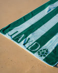 A green and blue striped beach towel laid diagonally on sandy ground, with the 'JAYD' printed in large letters along the bottom with the JAYD Gem icon near the corner.