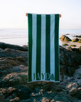 A person holds up a large beach towel with vertical green and white stripes on a rocky beach with the ocean in the background. The towel has the name 'JAYD' and a small JAYD Gem logo printed near the bottom.