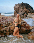 Model Chloe standing in front of a rock at the beach, wearing the mid-coverage JAYD Cali bottoms in black.