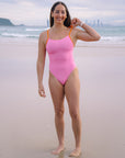 JAYD Swimwear athlete Jenaya Massie standing on a beach wearing the Airlie one piece swimsuit in Fairy Floss pink with contrasting Flare orange shoulder straps.