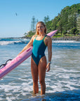 JAYD Swimwear model Brodee standing in shallow ocean water on a beach, wearing the Cove Finley one piece swimsuit with contrasting moss green shoulder straps, holding a pink ski.
