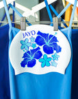 White swim cap hanging on a rack with blue hibiscus flowers, matching JAYD Swimwear's blue swimsuit range.