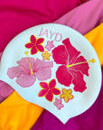 White Hibiscus Haven swim cap sitting on pink and orange JAYD one piece swimsuits, featuring pink and orange flowers.