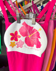 White swim cap with pink floral hibiscus design hanging on a rack with colourful pink and orange JAYD Swimwear one piece swimsuits.