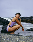 JAYD Swimwear model Sophie crouching on a rocky beach near the waters edge wearing the Ibiza one piece swimsuit in Lilac Haze with Acai hip cut-outs and shoulder straps.