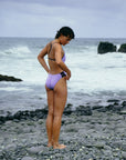 JAYD model Sophie wearing the purple Ibiza one piece swimsuit in lilac haze with bold contrasting dark purple acai across over shoulder straps for optimal support and thick chubby hip cut-outs, standing on a rocky beach with ocean waves in the background.