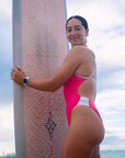 Athlete Jenaya Massie standing on the beach holding her surf board, ready to hit the waves in her JAYD one piece Ibiza swimsuit in vibrant pink sorbet with contrasting white hip cut-outs and back straps.