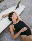 Model Sophie wearing the JAYD Isla one piece swimsuit, lying on the her surfboard on the beach.