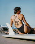 JAYD model Sophie sitting on a beach next to her surfboard in the black Isla one piece JAYD swimsuit with adjustable tie-back straps.