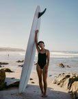 JAYD model Sophie wearing the Isla tie-back one piece swimsuit on a beach, posing next to her surfboard with waves in the distance.