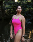 Athlete Jenaya standing in a river with a green forest behind her, wearing the JAYD Isla one piece in pink sorbet.