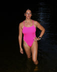 Bold pink sorbet Isla one piece modelled by athlete Jenaya Massi, standing in a iver at night surrounded by rippling water.