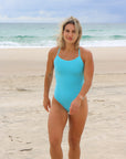 Front view of model walking on beach wearing Airlie One Piece Athletic Swimsuit in Mykonos Blue by JAYD Swimwear.