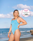 JAYD Swimwear model stands on a beach with the ocean and blue sky behind her, wearing the contrasting Waikiki blue Albi one-piece swimsuit with cancun straps. A circular text badge on the right reads 'Swim for a Cure' with all proceeds from this suit sale going to Cure Cancer.