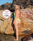A woman stands on a rocky beach wearing the JAYD Swimwear Zambia one piece in Kiwi green with contrasting tart pink shoulder straps and hip panels. A circular text badge on the right reads 'Swim for a Cure' with all proceeds from this suit sale going to Cure Cancer.