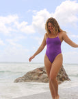 Female swimmer wearing a JAYD Swimwear athletic Lennox one piece swimsuit in Murasaki purple, walking along the beach.