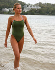 Front view of a female athlete walking on a beach while wearing a JAYD Swimwear one piece swimsuit, Lennox in Olive green with blue hip cut outs.