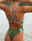 Back view of a female athlete on a beach wearing a JAYD Swimwear one piece swimsuit, Lennox in Olive green with blue hip cut outs.