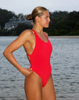 Front view of a woman in a JAYD Swimwear Levanto one piece swimsuit in Ruby, standing on a beach at sunrise.