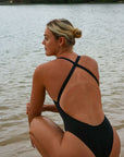 Back view of an athletic woman in a black JAYD Swimwear Levanto one piece swimsuit, sitting by the water at sunrise.