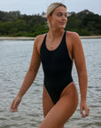 Front view of an athletic woman in a black JAYD Swimwear Levanto one piece swimsuit, walking on a beach at sunrise with water and trees in the background.