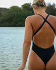 Back view of an athletic woman in a black JAYD Swimwear Levanto one piece swimsuit, walking into a body of water.