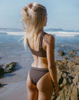 Woman in a brown JAYD Swimwear bikini top, Nalu in Cacao, standing on a rocky beach with ocean waves in the background