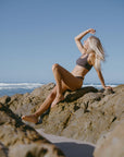Woman in a brown JAYD Swimwear athletic bikini top, Nalu in Cacao, sitting on a rock by the ocean with a clear blue sky.