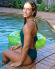 Model sitting by a pool wears JAYD Swimwear tie back athletic full coverage one piece: Riva in Cove