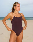 Front view of an athletic woman wearing a dark brown athletic JAYD Swimwear Riva one piece swimsuit in cacao, standing on a beach.