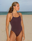 Front view of an athletic woman wearing a dark brown athletic JAYD Swimwear Riva one piece swimsuit in cacao, standing on a beach.