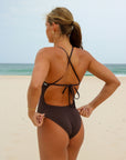 Close back strap view of an athletic woman wearing a dark brown athletic JAYD Swimwear Riva one piece swimsuit in cacao, standing on a beach.