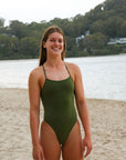 Front view of a female athlete wearing a JAYD Swimwear athletic one piece swimsuit, Riva in Olive green, standing on a beach.