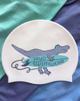 A white swim cap featuring a cartoon T-rex holding a teal surfboard with the text 'JAYD SWIMWEAR', displayed on a background of teal and navy blue fabric.