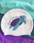 A white silicone swim cap featuring a custom printed design of a purple and blue sea turtle with a purple flower on its shell, alongside the text 'Jazzy Swimswear'. The swim cap is laid flat on a background of teal and purple fabric.