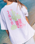 Woman wearing a white JAYD Swimwear oversized boxy tee with a pink cowgirl boots and cactus graphic print on the back, styled at the beach.