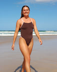 Australian triathlete, Charlotte Derbyshire, in a Cacao Brown JAYD Swimwear athletic Zambia one piece swimsuit, walking on a sandy beach with clear blue sky.
