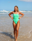 Front view of a female swimmer in a Meadow green athletic JAYD Swimwear Zambia one piece swimsuit walking on a beach with ocean waves in the background