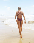 Back view of surf Ironwoman, Brodee Trinca, walking on a beach wearing JAYD Swimwear athletic one piece swimsuit, Apsley in Sangria.