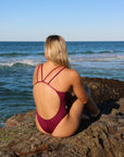 Back view of model sitting on a rock wearing JAYD Swimwear athletic one piece Finley in Sangria.