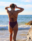 Back view of model walking into the water wearing JAYD Swimwear athletic tie-back one piece Riva in Sangria.