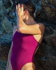Side view of model leaning against a rock wearing JAYD Swimwear athletic tie-back one piece Riva in Sangria.